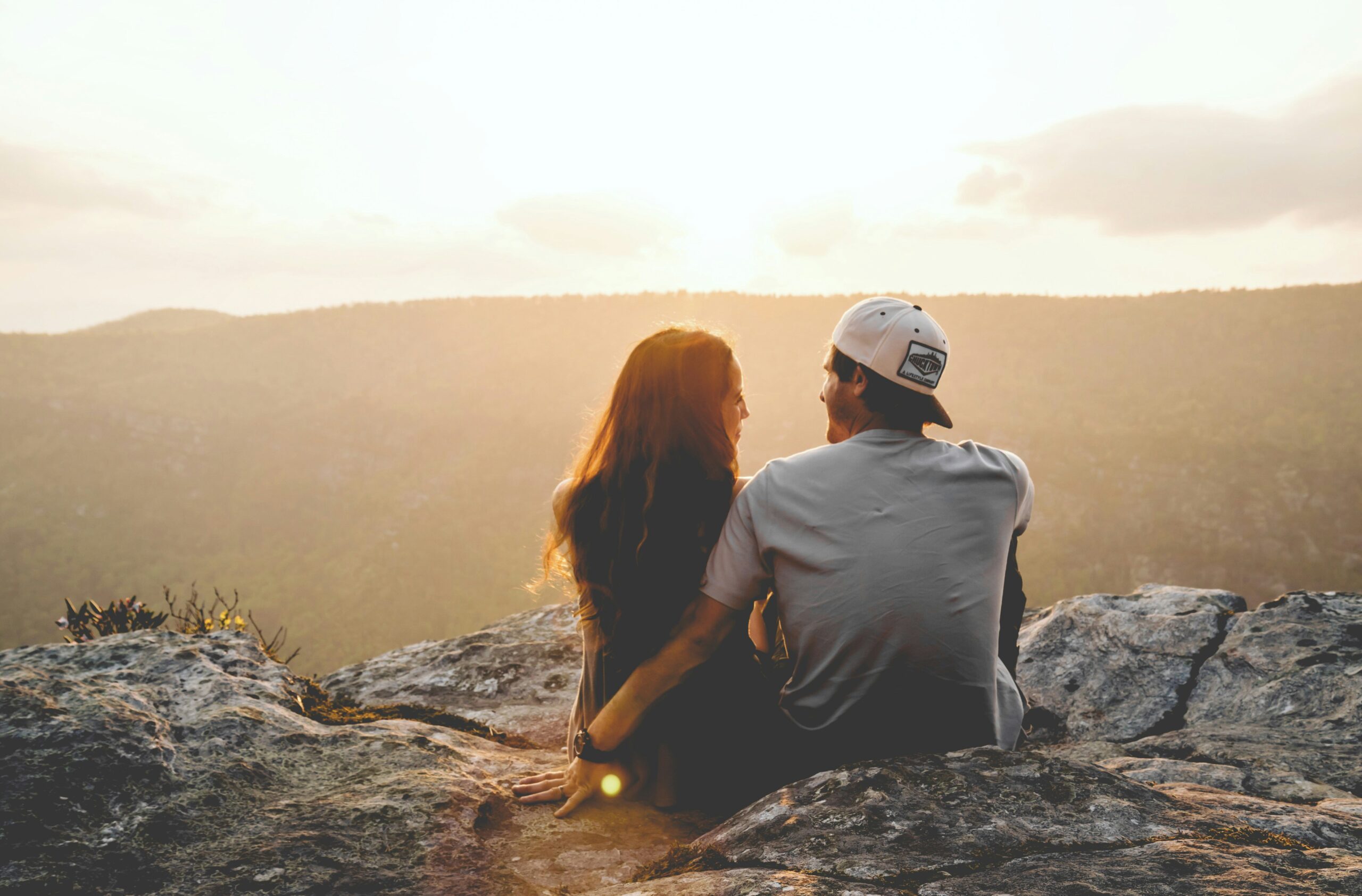 A couple sits close together on a rocky overlook, bathed in golden light, gazing at each other as the sun sets over the hills.
