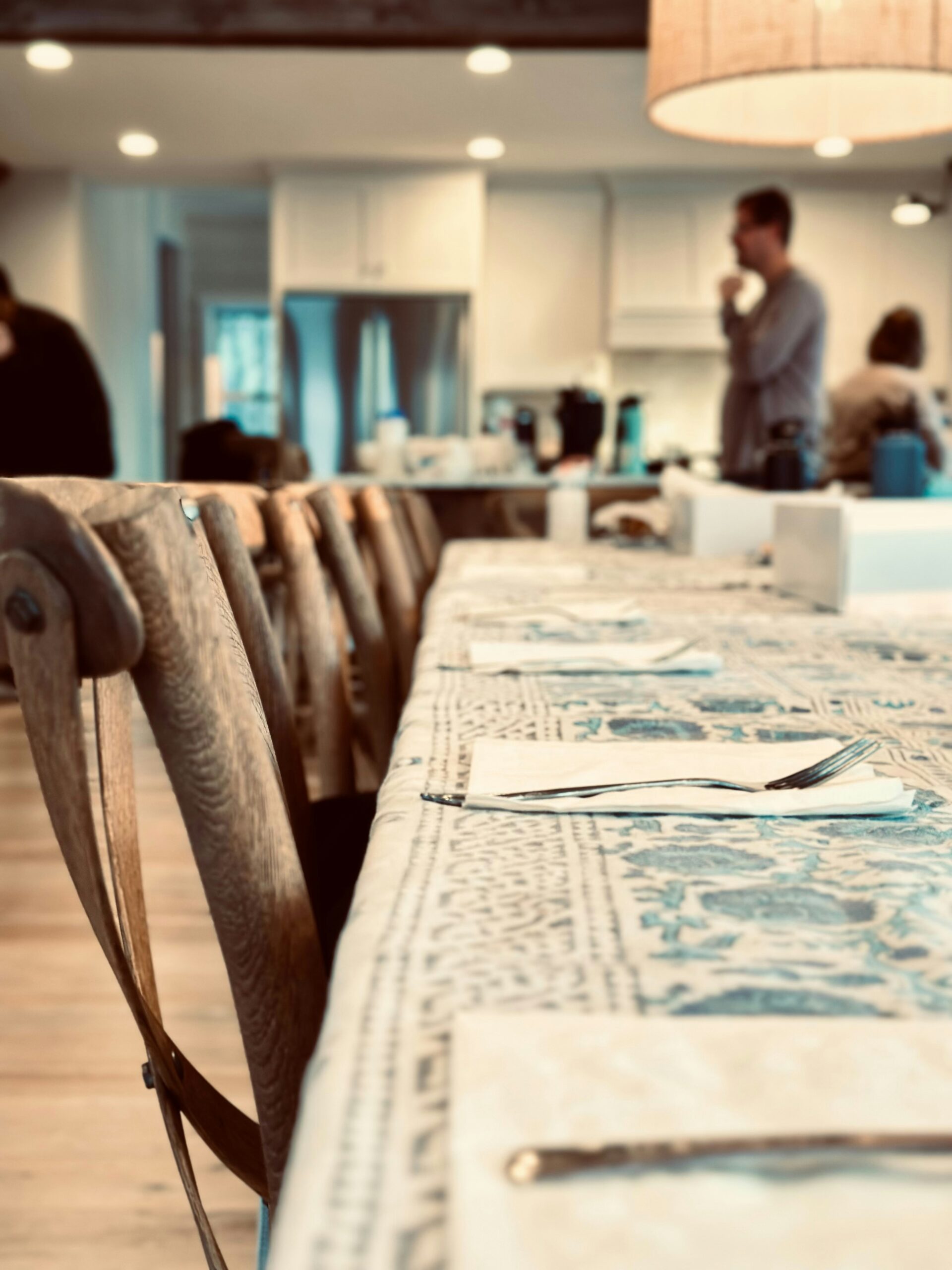 A long dining table set with napkins and cutlery, with people gathered in the blurred background of a warm kitchen during a family gathering.
