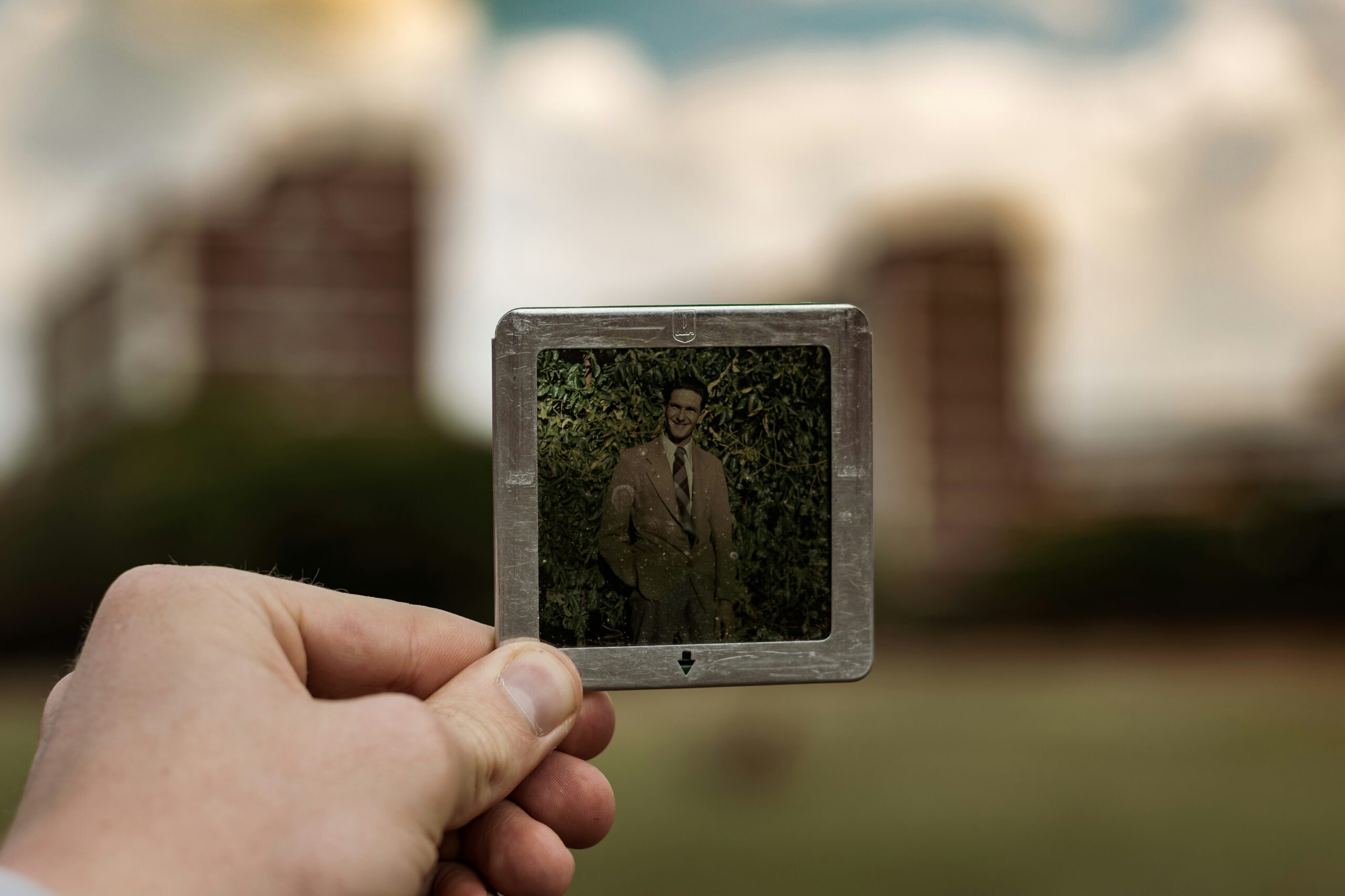 A hand holding an old photograph in front of a blurred house, representing memory, grief, and continuing bonds after the death of a loved one.